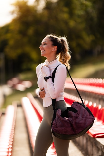Athletische Frau mit einem lila Sporttasche und einem weißen Oberteil, die auf einer Tribüne steht und lächelt.